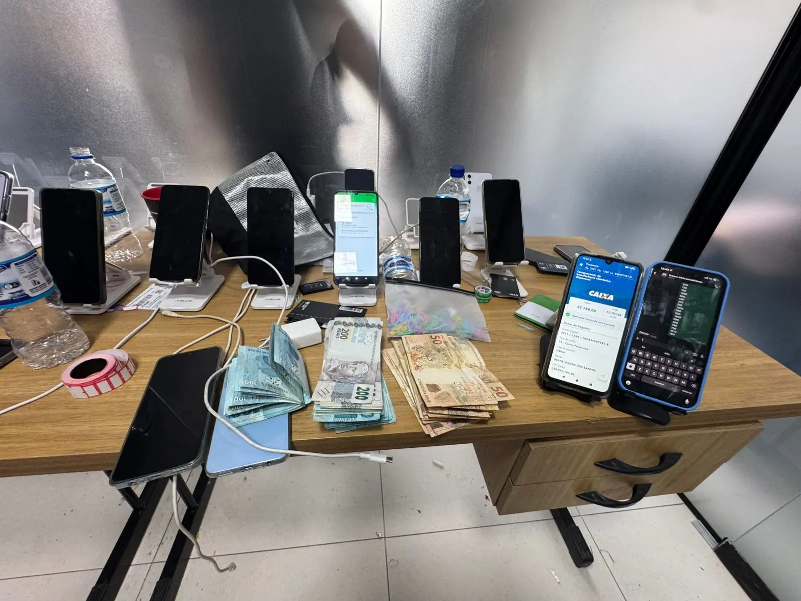 Mesa com dezenas de celulares em suportes, cartões bancários e maços de dinheiro apreendidos pela Polícia Civil